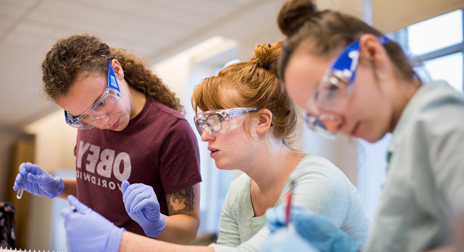 Photo of three female Chatham University biochemistry students wearing blue gloves and goggles, working in a lab holding glass test tubes. 